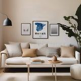 Spain map keepsake collage in modern black frame hung on living room wall above an ivory coloured couch with neutral coloured pillows. A tall plant is to the right and a small table in front.