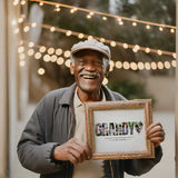 Older man wearing hat, holding grandy photo collage in frame, while standing outside with string lights in the background.