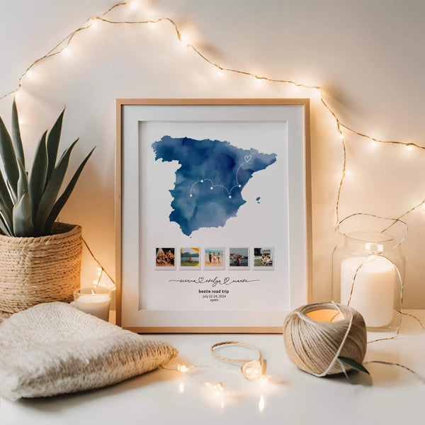 Spain map keepsake collage in a sleek wood frame stands between a potted plant and a white candle. Lit string lights surround the scene.